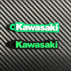 以下が含まれることがあります： Kawasakiのロゴが入った2つの長方形のラバーキーホルダー。上のキーホルダーは鮮やかな緑色のベースに白い文字、下のキーホルダーは黒いベースに鮮やかな緑色の文字がデザインされています。各ピースの左側には円形の穴が開いています。