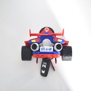 May include: A red, blue, and black toy race car with the text "LETS-A-GO!" on the back. The car has large black tires and a remote control hanging below. The car has a Mario logo on the top.