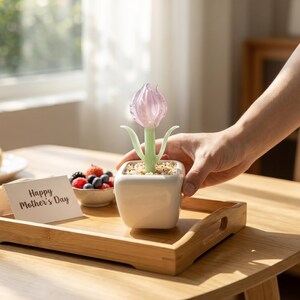 Op de afbeelding: Een glazen tulp in een witte vierkante pot, op een houten dienblad. De tulp heeft een roze bloem en groene stengel en bladeren. Een klein kaartje met de tekst "Happy Mother's Day" is zichtbaar.