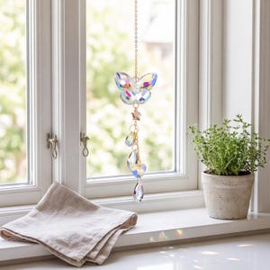 May include: A hanging suncatcher ornament featuring a faceted iridescent butterfly crystal suspended from a gold-toned metal chain, accented with smaller star-shaped and teardrop-shaped crystals that catch and reflect light in various colors.