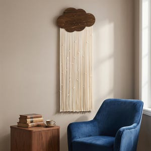 May include: A wooden cloud-shaped wall hanging with a dark brown finish. Below, long cream-colored cords hang down, knotted at intervals. The wall decor is mounted on a neutral-colored wall, creating a minimalist aesthetic.