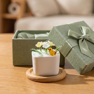 Könnte beinhalten: Ein kleiner weißer quadratischer Pflanztopf mit Glasblumen und einer gelben Vogelfigur, auf einem Holzuntersetzer. Eine grüne Geschenkbox mit Schleife befindet sich im Hintergrund. Das dekorative Miniaturgarten-Objekt.