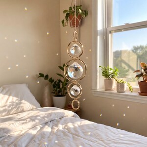 Puede incluir: Un atrapasoles colgante con tres esferas de cristal transparente facetado suspendidas dentro de anillos metálicos dorados interconectados. El adorno cuelga de una cadena delicada y captura la luz solar para proyectar reflejos.