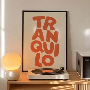 May include: A white record player with a black vinyl record is on a wooden surface. A framed poster with the word "TRANQUILO" in orange letters is behind the record player. A round, white and yellow lamp is to the left.
