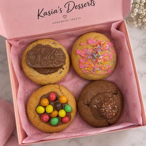May include: A pink gift box labelled Kasia's Desserts containing four cookies. One cookie has a chocolate frog, one has pink icing with sprinkles, one has colourful candy-coated chocolates, and one has a hazelnut chocolate truffle on top.
