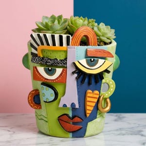 Puede incluir: Una colorida maceta de cerámica con forma de rostro, con detalles en verde, azul, naranja y morado. La maceta presenta un diseño caprichoso con rasgos faciales abstractos y contiene varias suculentas verdes. El fondo está dividido entre rosa y azul.