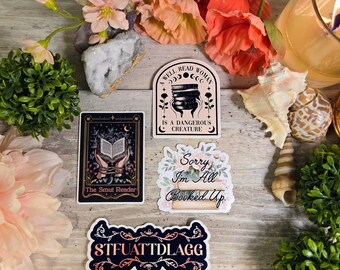 May include: A collection of stickers with various designs and text, including "The Smut Reader," "Sorry I'm All Booked Up," and "STFUATTDLAGG." The stickers are arranged with flowers, shells, and a lit candle on a wooden surface.
