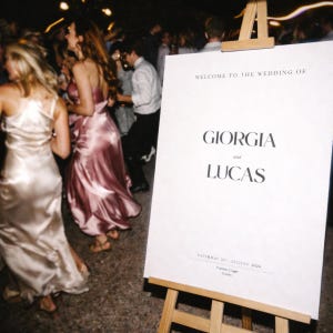 Op de afbeelding: Een trouwreceptie met een bord met de tekst "Welkom bij de bruiloft van Giorgia en Lucas." Het bord staat op een ezel. Gasten dansen op de achtergrond, met twee vrouwen in satijnen jurken.