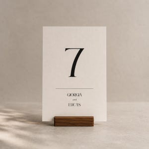 Op de afbeelding: Een cr&egrave;mekleurige tafelnummerkaart met het nummer 7 in zwart, en de namen "Giorgia en Lucas" eronder. De kaart wordt vastgehouden in een donkerbruine houten houder. Het ontwerp is minimalistisch en elegant.