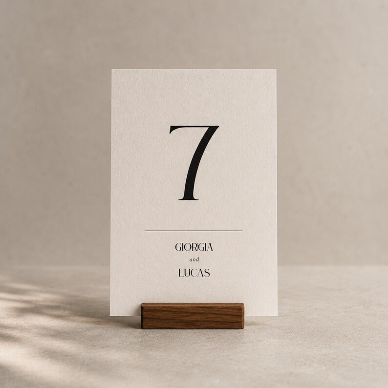 Op de afbeelding: Een cr&egrave;mekleurige tafelnummerkaart met het nummer 7 in zwart, en de namen "Giorgia en Lucas" eronder. De kaart wordt vastgehouden in een donkerbruine houten houder. Het ontwerp is minimalistisch en elegant.