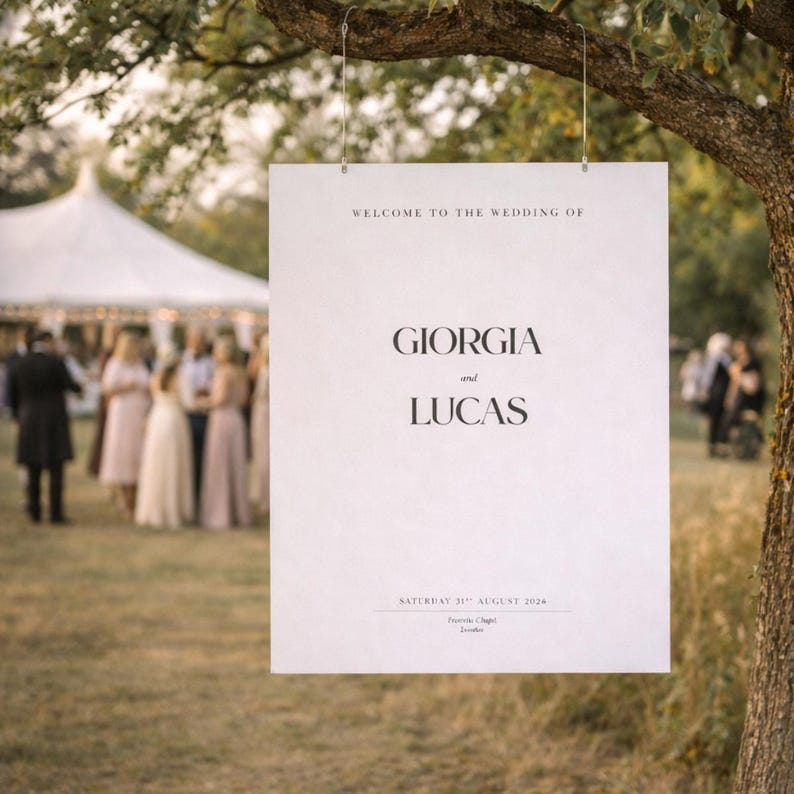 Op de afbeelding: Een wit trouwbord hangt aan een tak. Het bord leest "WELCOME TO THE WEDDING OF GIORGIA and LUCAS" met datum en locatie eronder. Een bruiloftgezelschap en tent zijn zichtbaar op de achtergrond.