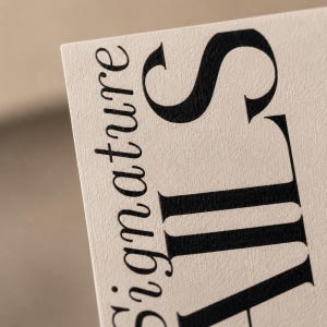 Op de afbeelding: Close-up van een beige visitekaartje met het woord "signature" in een zwart, elegant lettertype. Het woord "signature" is verticaal geori&euml;nteerd, met de letter "S" prominent weergegeven.