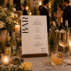 Op de afbeelding: Een drankmenu op een houten standaard, met het woord "BAR" in grote, zwarte letters. Het menu bevat wijn, bier, sterke drank en frisdranken. De tafel is gedekt met drankjes, olijven, citroenen en kaarsen.