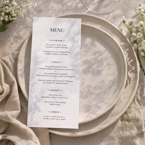 Könnte beinhalten: Eine gedruckte Menükarte mit dem Wort „MENU“ oben, die Vorspeisen, Hauptgerichte, Desserts und Käseoptionen auflistet. Die Karte liegt auf einem weißen Teller, mit einem zweiten Teller darunter. Weiße Blumen im Hintergrund.