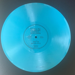 May include: A translucent bright blue vinyl record with a central circular label. The label features black text reading Dark Horse, Paul, with Buddy Holly Fa-la-la-la-la... La-la-la-faaaashh, SFF 32 (an audio issue), and 33 1/3 RPM MONAURAL.