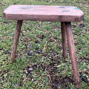 May include: A rustic wooden stool with three legs and a rectangular top. The stool is painted a light pink color with some green paint accents. The stool is sitting on a bed of green grass.