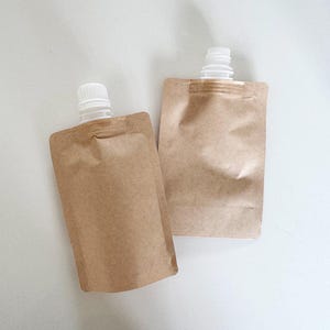 Puede incluir: Dos bolsas de papel kraft marrón con boquillas de plástico blanco. Las bolsas son rectangulares con esquinas redondeadas y están sobre una superficie blanca. Las boquillas están en la parte superior de las bolsas.