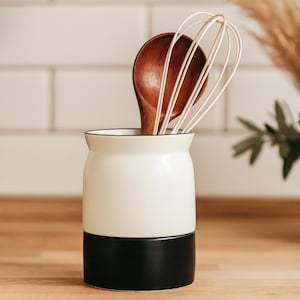 Op de afbeelding: Een tweekleurige keukengerei houder in crème en zwart, met een houten lepel en een garde. De houder staat op een houten oppervlak. Het bovenste deel is crème, het onderste deel is zwart.