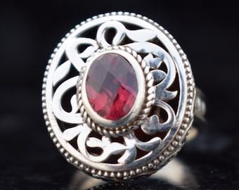 Puede incluir: Anillo de plata con una gema roja ovalada y facetada en el centro. El anillo tiene un diseño calado ornamental alrededor de la gema y un borde con cuentas. El anillo se muestra sobre una superficie reflectante.