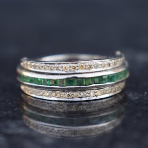 Op de afbeelding: Een zilveren ring met drie banden. De bovenste en onderste banden zijn versierd met kleine, heldere stenen, terwijl de middelste band een rij vierkante, groene stenen heeft. De ring staat tegen een donkere achtergrond.