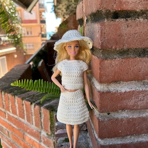 May include: A doll wearing a cream-colored, crocheted outfit, including a crop top, skirt, and sun hat. The doll has blonde hair and is posed outdoors against a brick wall. The outfit appears handmade and has a textured, open weave.