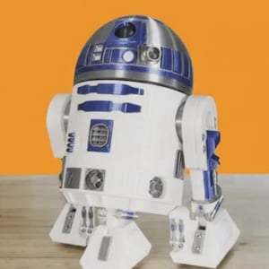 Può includere: Una figura di droide R2-D2 bianca e blu con una testa a cupola argentata. Il personaggio di Star Wars ha un corpo rettangolare con dettagli e accenti blu. Il droide è in piedi su una superficie di legno contro uno sfondo arancione.