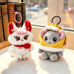 May include: Two plush keychains. One is a white fox with red ears, a red bow, and a pink beaded chain. The other is a gray bear wearing a yellow hooded cape with a small crown, attached to a black metal ring. Both have embroidered facial features.