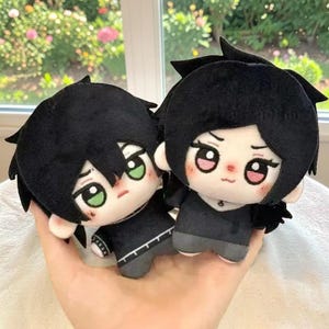 以下が含まれることがあります： 黒い髪と黒い服を着た2体のぬいぐるみ。左側のぬいぐるみは緑色の目で無表情、右側のぬいぐるみはピンク色の目で小さな口をしています。どちらのぬいぐるみも柔らかい布製で、顔のパーツは刺繍で表現されています。