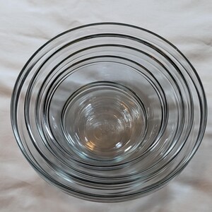May include: A set of five clear glass mixing bowls nested together. The bowls are of varying sizes, with a simple, rounded design. The bowls are arranged on a white surface.