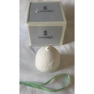 May include: A white porcelain bell with a detailed relief design, a small loop at the top, and a green ribbon. The bell is next to a box with the "LLADRÓ" logo.