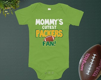 Może przedstawiać: Limonkowy body niemowlęce z napisem "MOMMY'S CUTEST PACKERS FAN!" w kolorze białym, zielonym i pomarańczowym. Grafika piłki nożnej znajduje się na środku. Brązowa piłka nożna po prawej stronie.
