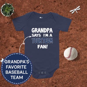 May include: A navy blue baby onesie with white text that reads "Grandpa says I'm a your team fan!"  A white arrow points to the text.  The onesie has a white baseball graphic on the front.  The text "Grandpa's favorite baseball team" is in a white circle with a dashed border.