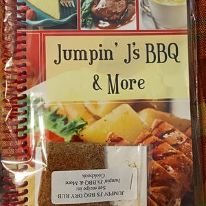 May include: A spiral-bound cookbook titled Jumpin' J's BBQ & More by Jason Wiggs, featuring food photography of cake, steak, and soup. A small clear plastic bag containing brown dry rub seasoning is attached to the front cover with a white label.