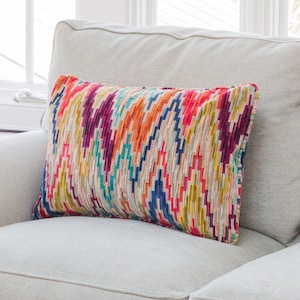 May include: A living room scene featuring a light gray sofa with a colorful patterned pillow. A blue and white ceramic lamp with a cream shade sits on a dark wooden side table. A red-brown coffee table is in the foreground.