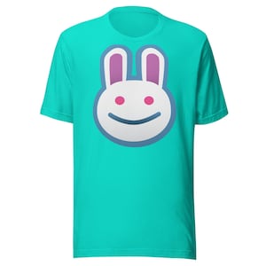 Puede incluir: Una camiseta turquesa con la cara de un conejito de dibujos animados. El conejo tiene pelaje blanco, ojos rosas y orejas moradas, delineadas en azul. La boca del conejo es una línea azul curva. La camiseta es de material suave.