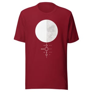 Puede incluir: Una camiseta de manga corta de color rojo oscuro con un gráfico de una luna dividida en una mitad blanca sólida y una mitad gris texturizada. Debajo de la luna hay una línea vertical con pequeños círculos y cuadrados, junto a etiquetas de texto blancas.