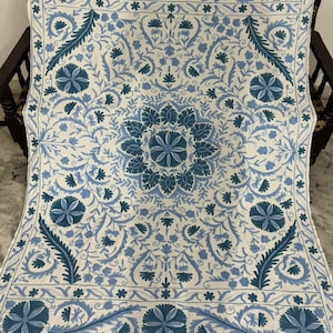 May include: A white and blue floral embroidered throw blanket. The blanket features a detailed pattern of flowers and leaves in shades of blue, with a border of smaller floral designs. The blanket is draped over a wooden chair.