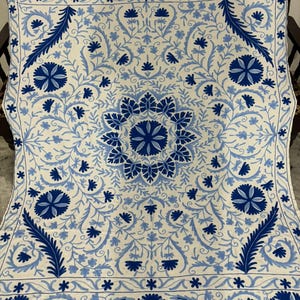 May include: A white and blue embroidered textile featuring a floral pattern. The design includes large, stylized flowers, leaves, and vines in shades of blue against a white background. The textile is likely a decorative item, such as a throw or wall hanging.