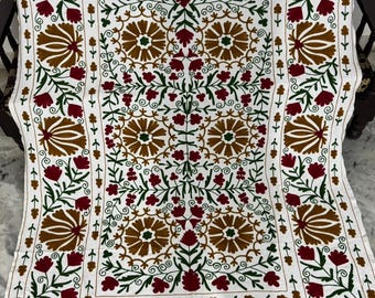 May include: White textile with an embroidered floral pattern. The design features brown, red, and green flowers and leaves. The embroidery is detailed and covers the entire surface, creating a decorative and textured appearance. The textile is likely a tablecloth or a decorative throw.