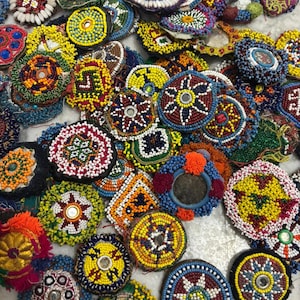 Puede incluir: Una colección de adornos de cuentas coloridos y hechos a mano. El surtido incluye varias formas y tamaños, con patrones intrincados en tonos de rojo, amarillo, azul y blanco. Algunas piezas tienen espejos o borlas.