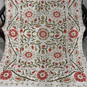 Puede incluir: Textil blanco con intrincados bordados con patrones florales rojos y marrones. El diseño incluye grandes flores estilizadas, hojas y vides, creando una composición simétrica y detallada. Los bordes están acabados con un delicado borde.