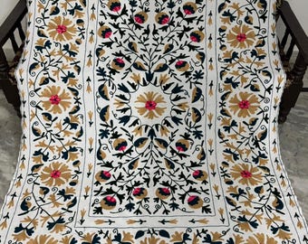 May include: White embroidered textile featuring a floral design with gold, pink, and navy blue accents. The intricate pattern includes stylized flowers and leaves, creating a decorative and detailed look. The textile is draped over a wooden chair.
