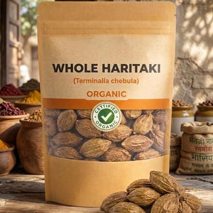 May include: A tan paper bag labeled "WHOLE HARITAKI (Terminalia chebula) ORGANIC" with a clear window showing brown, oval-shaped seeds. A "CERTIFIED ORGANIC" sticker is visible. A small pile of the seeds sits in front of the bag.