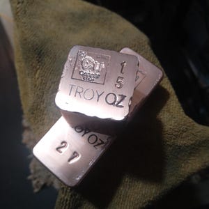 May include: Three copper bars with stamped markings, including the element symbol Cu, the text "Copper", and "TROY OZ". One bar is marked "1.5 TROY OZ" and another "2.7 TROY OZ". The bars have a rough, textured surface and a reddish-brown colour.