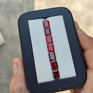 Puede incluir: Una colección de siete gemas rectangulares de color rojo intenso dispuestas verticalmente sobre un fondo blanco dentro de un estuche negro. Las gemas tienen una superficie pulida y reflectante, mostrando su rico color. El estuche se sostiene en una mano, con un fondo neutro.
