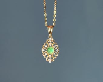 Puede incluir: Un collar dorado con un colgante con una piedra preciosa verde en el centro, rodeada de pequeñas piedras preciosas transparentes. El colgante tiene un diseño calado y ornamentado. La cadena es delicada y también dorada.