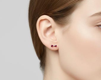 Puede incluir: Primer plano de una oreja con pendientes de botón en forma de lazo. Cada pendiente tiene una gema de color rojo oscuro en el centro. Los pendientes están engarzados en un metal claro, posiblemente plata u oro blanco.