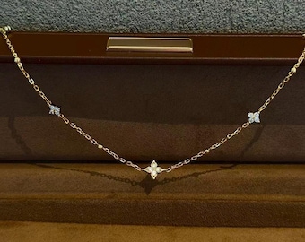 Puede incluir: Un delicado collar de oro con diseños florales de cuatro pétalos y pequeños detalles brillantes. El collar se exhibe dentro de un joyero marrón, mostrando su elegante diseño y artesanía.