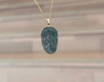 Puede incluir: Un collar dorado con un colgante en forma de hoja. El colgante es de color verde apagado con vetas detalladas, engastado en un marco dorado. La cadena es delicada y cuelga del colgante.