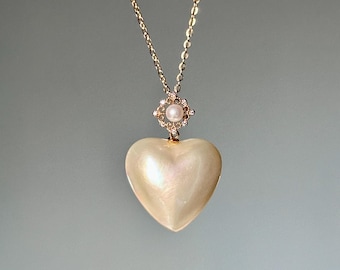 Puede incluir: Un collar dorado con un colgante en forma de corazón de color crema nacarado. La delicada cadena dorada sostiene el colgante, con una pequeña perla engastada en un engaste en forma de diamante sobre el corazón. Una joya romántica.
